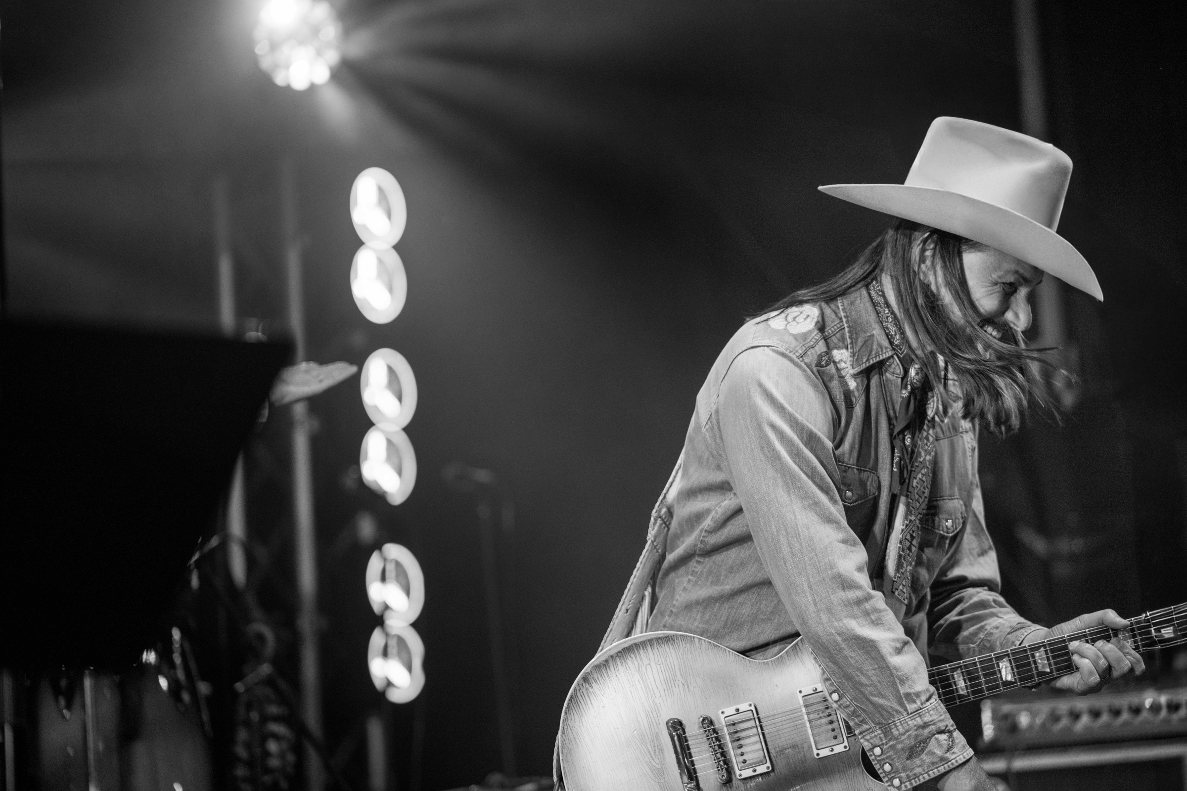 Duane Betts playing guitar on stage in a white hat, stage lights behind him, in black and white