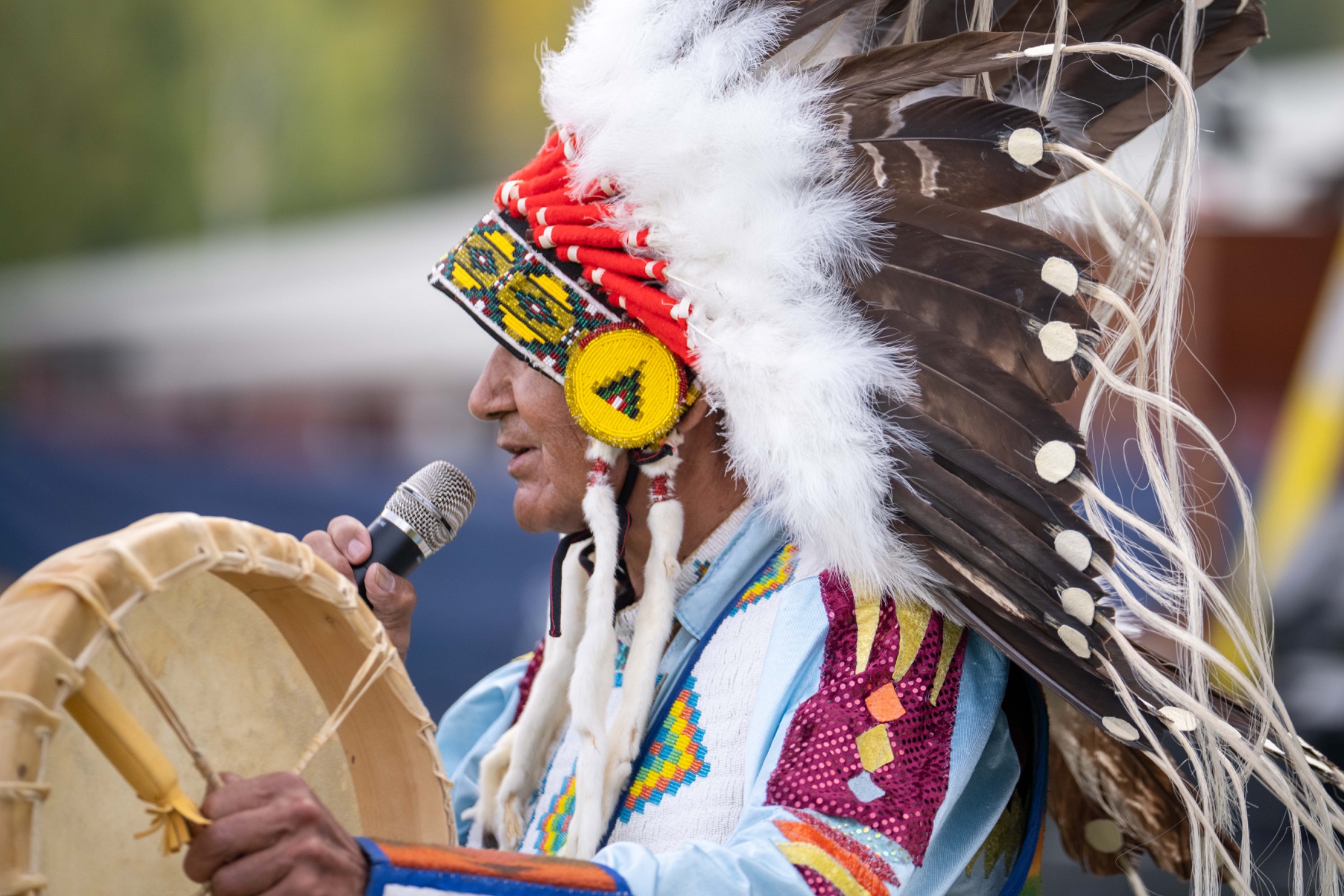An elder in an eagle-feather warbonnet and beaded regalia singing into a microphone at Horseshoe, holding a hand drum