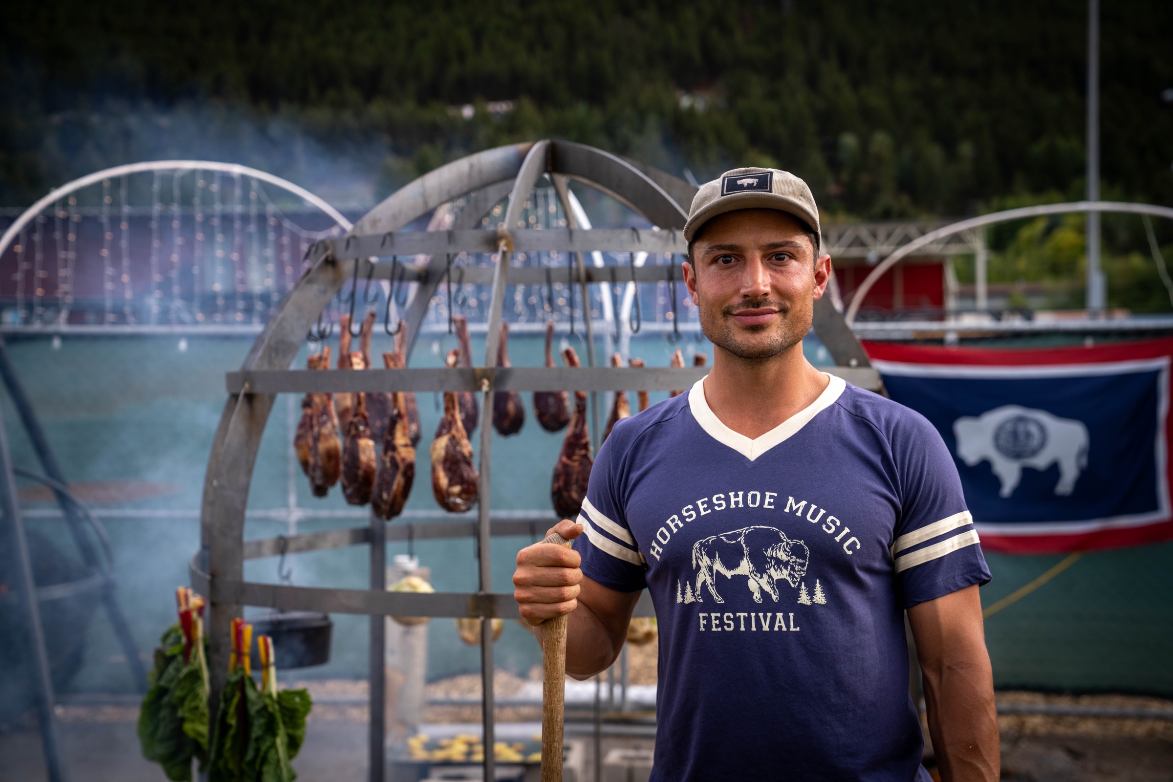 A cook in a Horseshoe Music Festival shirt standing at the smoking rack, cuts of meat hanging behind him in the smoke